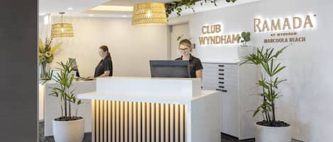Un mostrador de recepción de diseño moderno, con un área blanca, dos plantas en macetas y un cartel que dice "CLUB WYNDHAM RAMADÁ MARCOOLA BEACH".