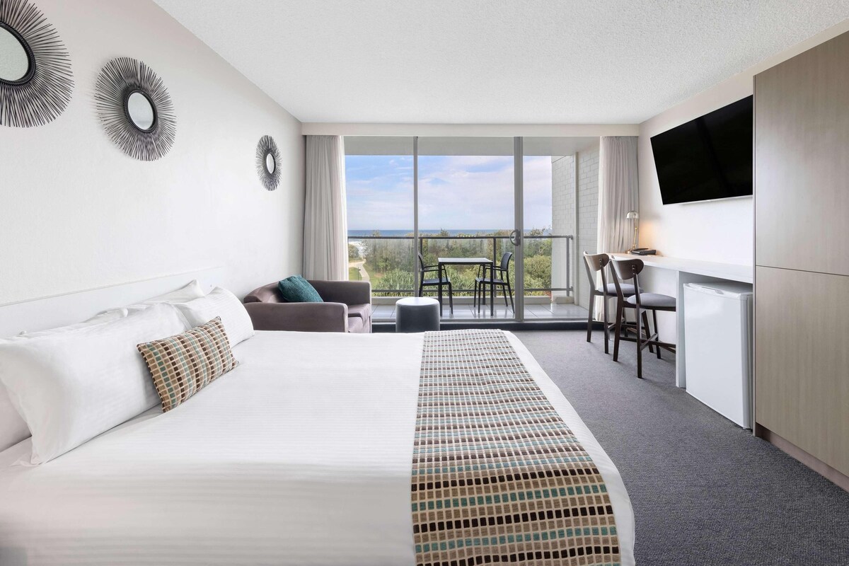 Habitación de hotel con una cama grande, un escritorio con sillas, un televisor y un balcón con vistas a la playa.