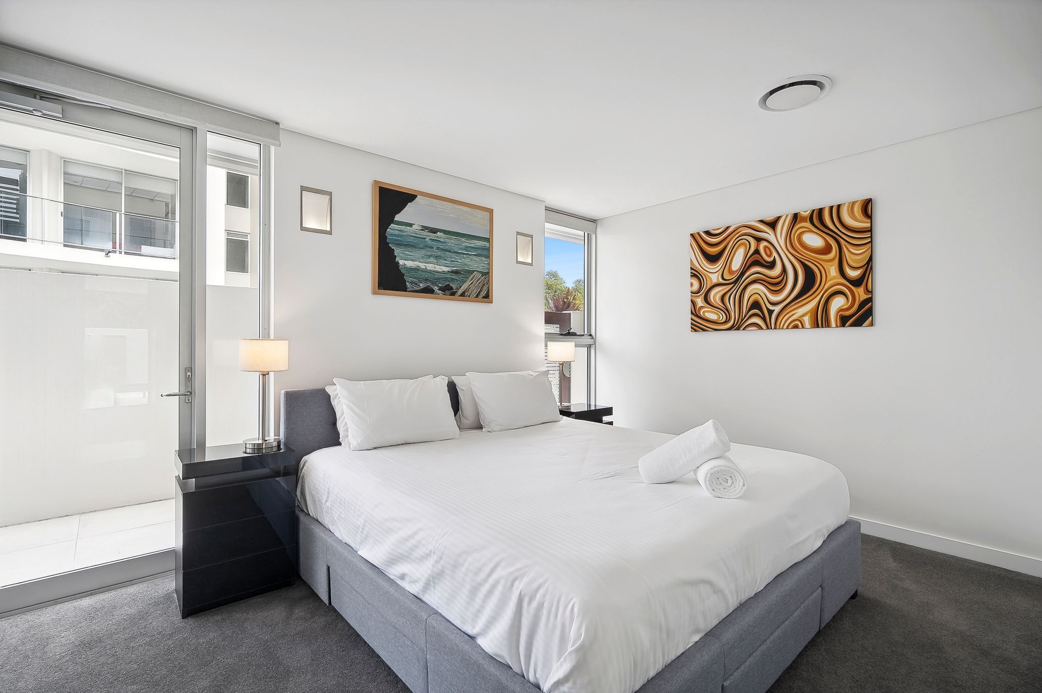 Una habitación de hotel moderna con una cama grande, un ventanal de vidrio que da a un balcón y dos obras de arte en las paredes.
