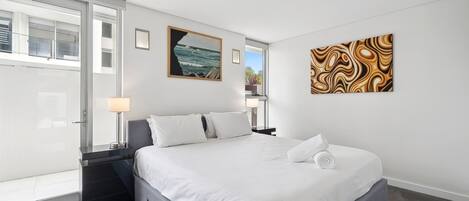 Una habitación de hotel moderna con una cama grande, un ventanal de vidrio que da a un balcón y dos obras de arte en las paredes.