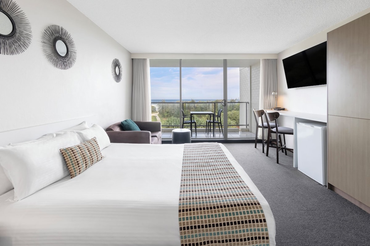 Habitación de hotel con cama, escritorio, silla, televisión y balcón con vistas.
