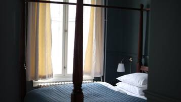 A four-poster bed with a quilted blue bedspread, a bedside lamp, and a window with sheer curtains.