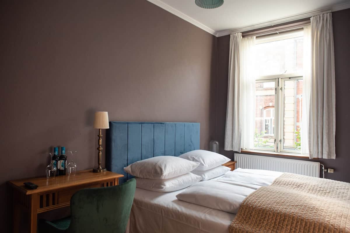 A hotel room with a bed, a bedside table with a lamp, a chair, and a window with curtains.