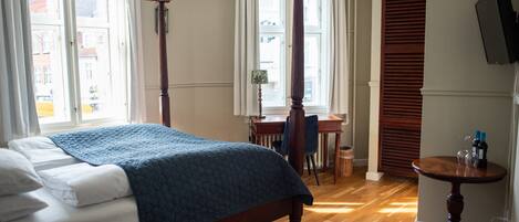A four-poster bed with a blue quilt, wooden flooring, a small table with bottles, and a TV on the wall.