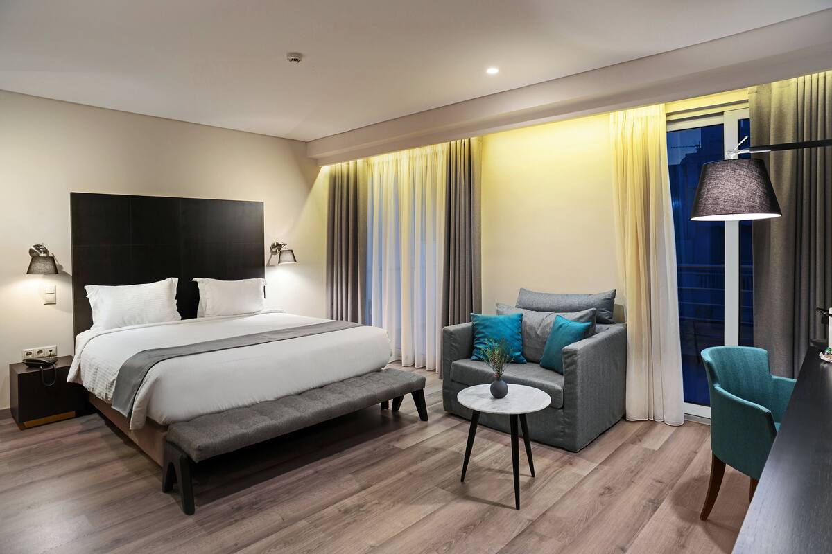 Quarto de hotel moderno com uma cama grande, um sofá cinzento, uma pequena mesa redonda e uma cadeira.