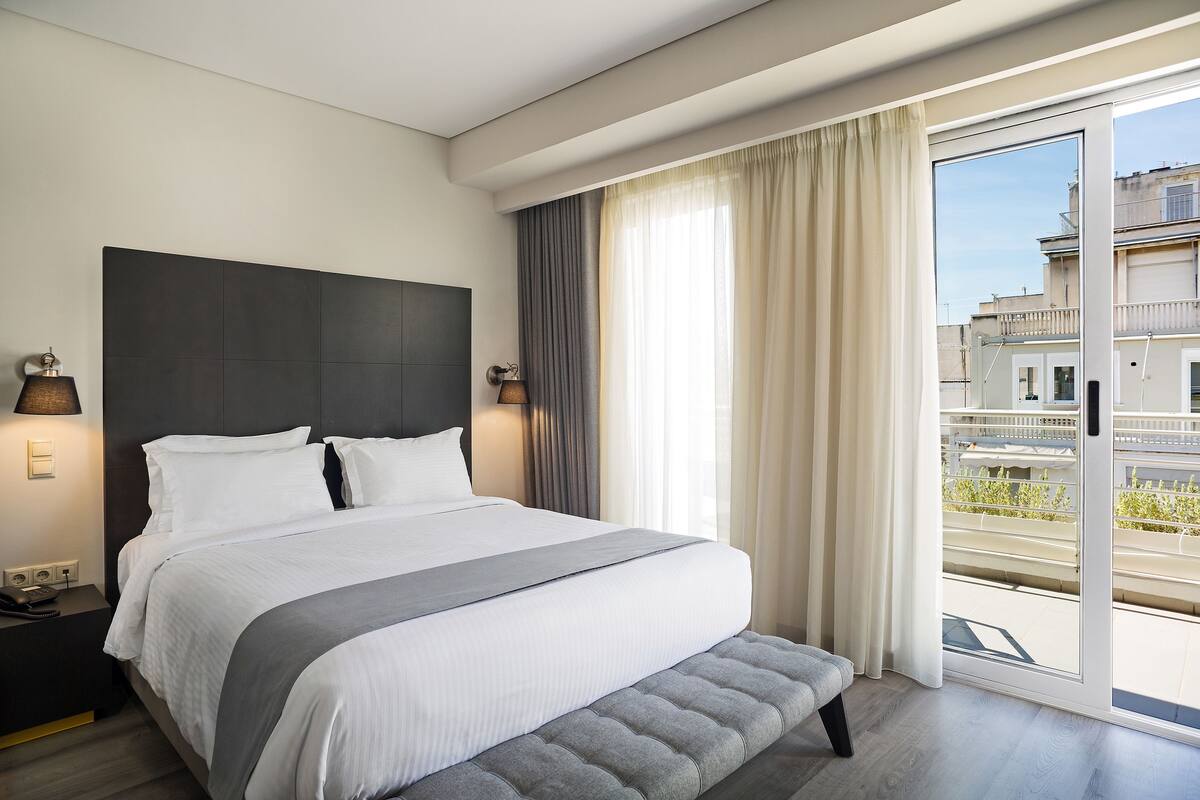 Quarto de hotel moderno com uma cama grande, um banco ao pé, uma mesa de cabeceira com luminária e vista para a cidade através de uma janela ampla.