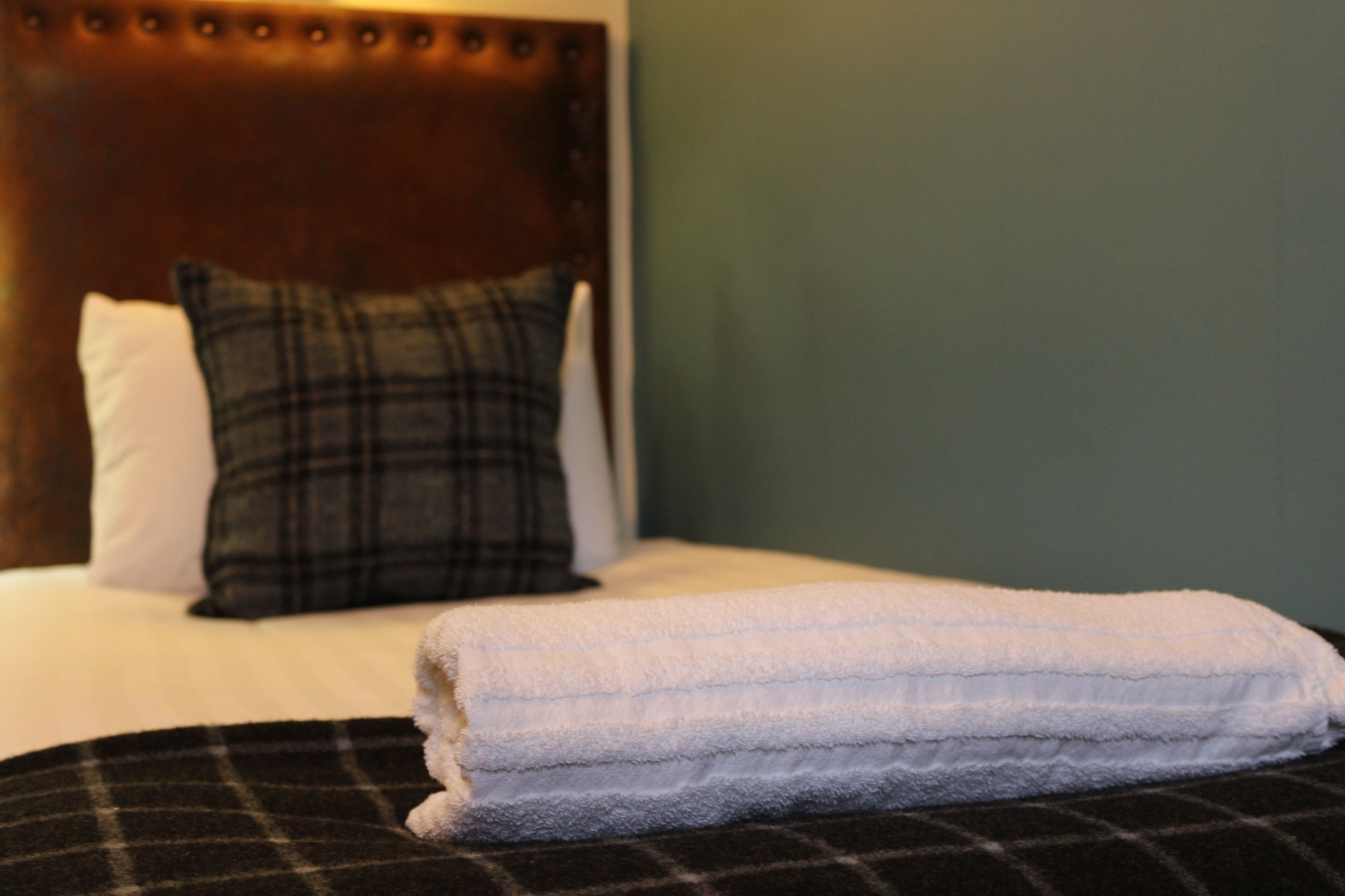 A neatly made bed with a white pillow and a plaid pillow, a folded white towel, and a dark headboard.