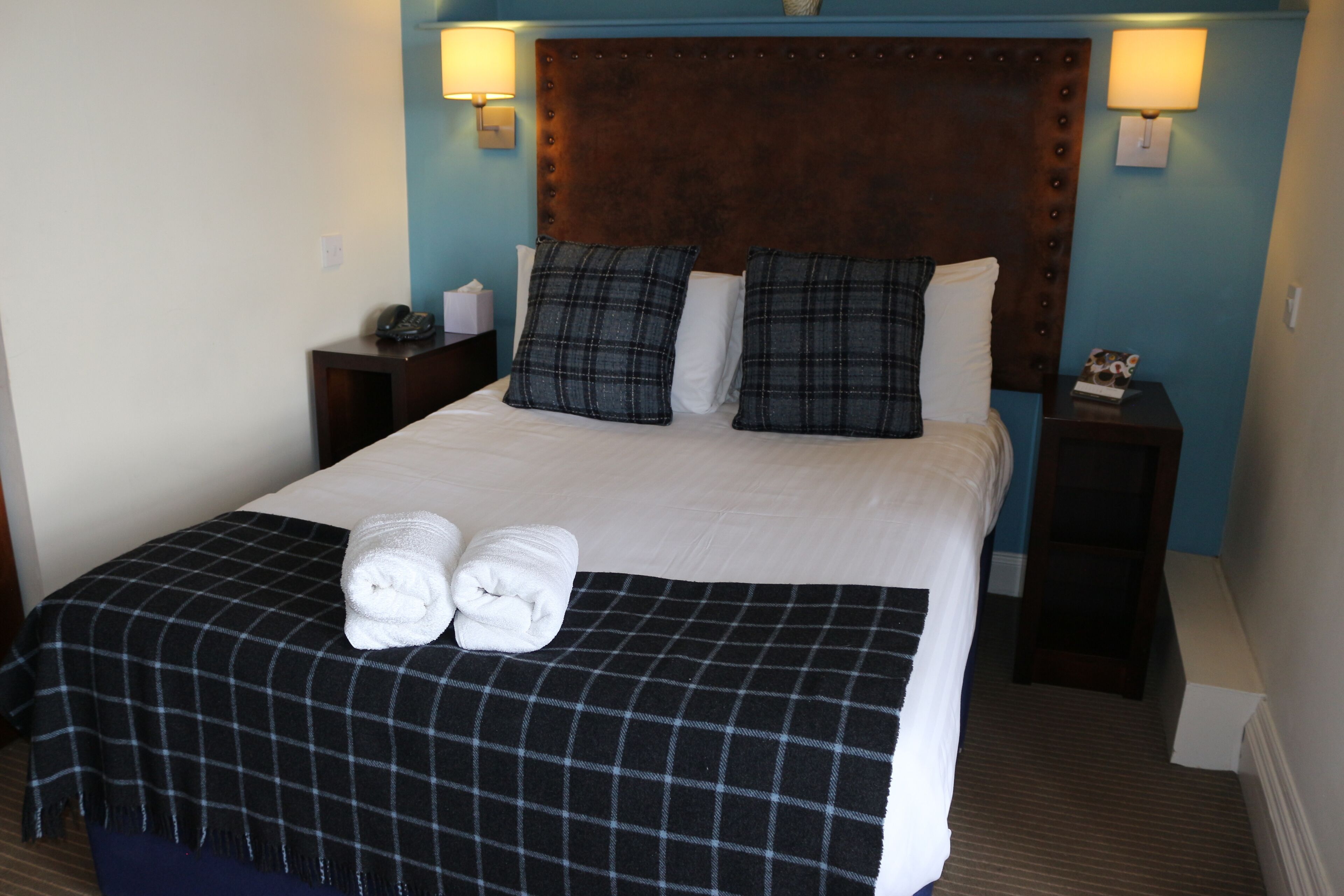 A neatly made bed with a checkered bedspread, two pillows, and two rolled towels. There are two bedside tables with lamps and a small decorative item on one.