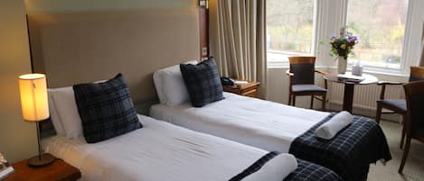 A hotel room with two beds, a nightstand with a lamp, a small table with a vase of flowers, and a chair.