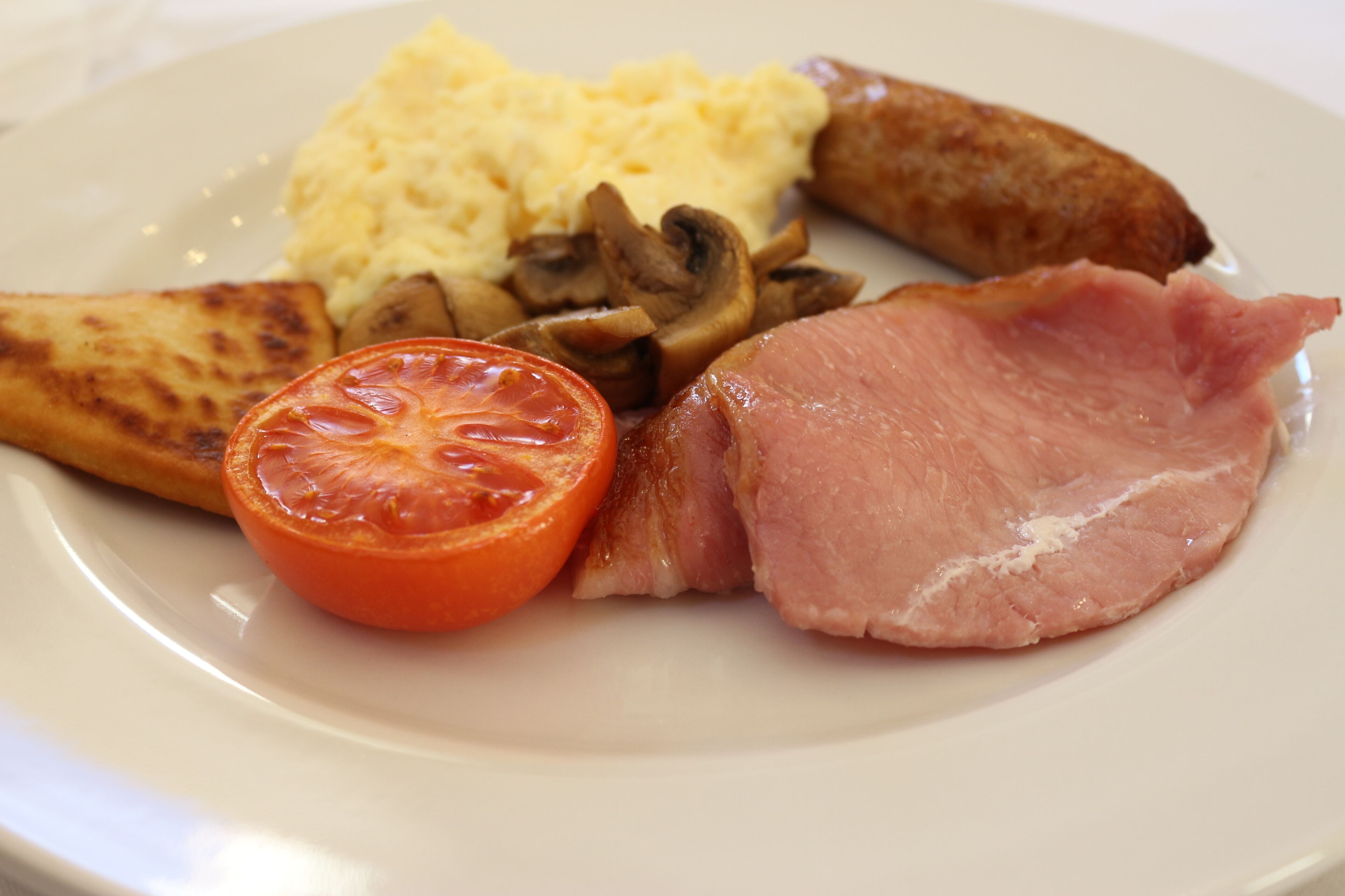 A plate with scrambled eggs, grilled mushrooms, a slice of ham, a grilled tomato, and a piece of toast.