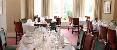 Un comedor con mesas redondas dispuestas para varios invitados, con manteles blancos, cubertería ordenada y sillas con tapicería verde.