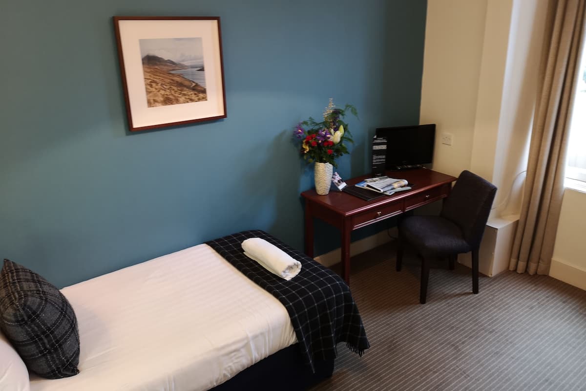 Une chambre d’hôtel avec un lit, un bureau avec un ordinateur, une chaise, un vase de fleurs et un tableau encadré au mur.