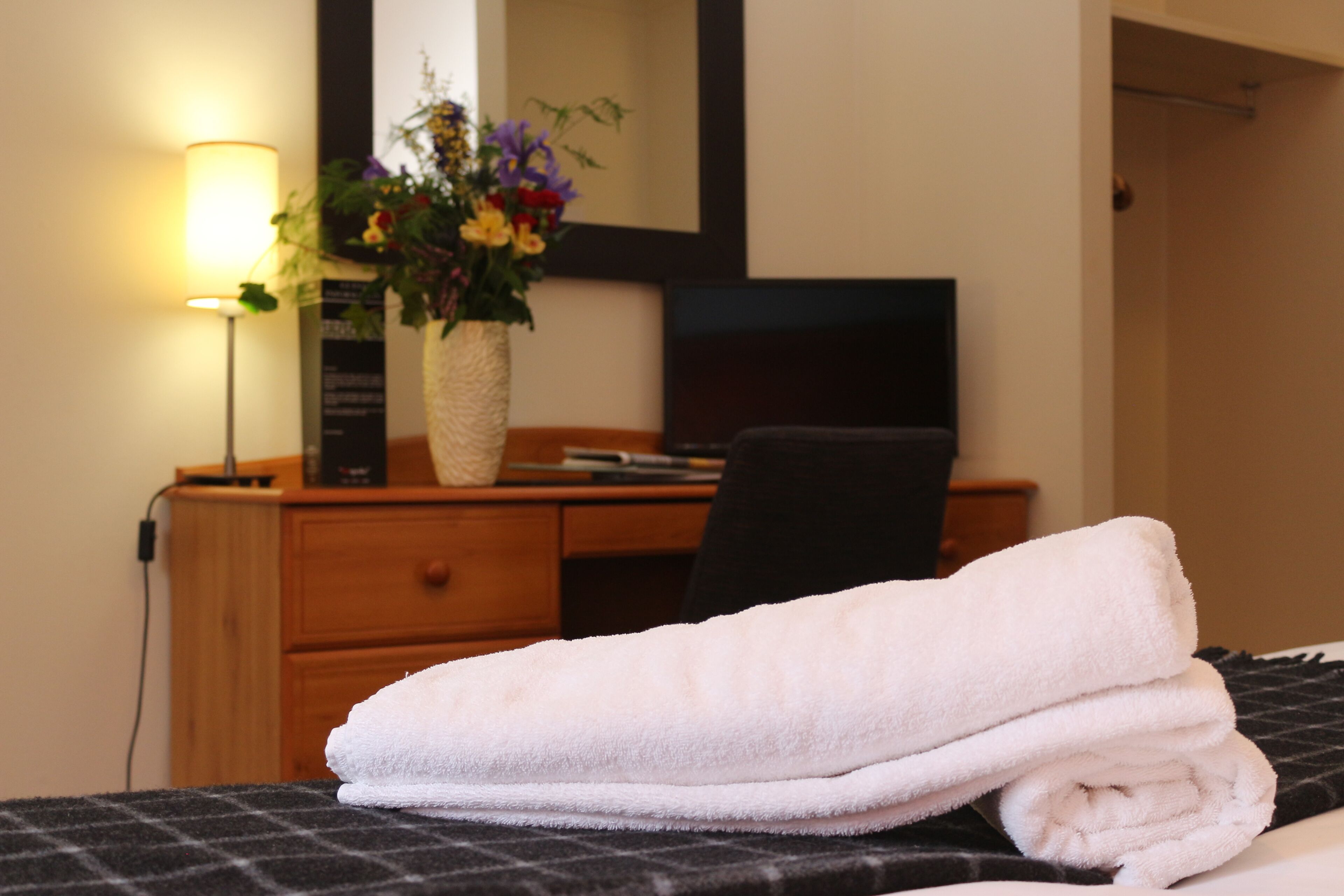 A neatly made bed with a folded white towel, a bedside lamp, a wooden dresser, and a vase with flowers.