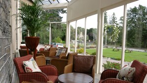 A sunlit patio with wicker furniture, a potted plant, and a view of a green landscape through large windows. - Craiglynne Hotel (Grantown-on-Spey)