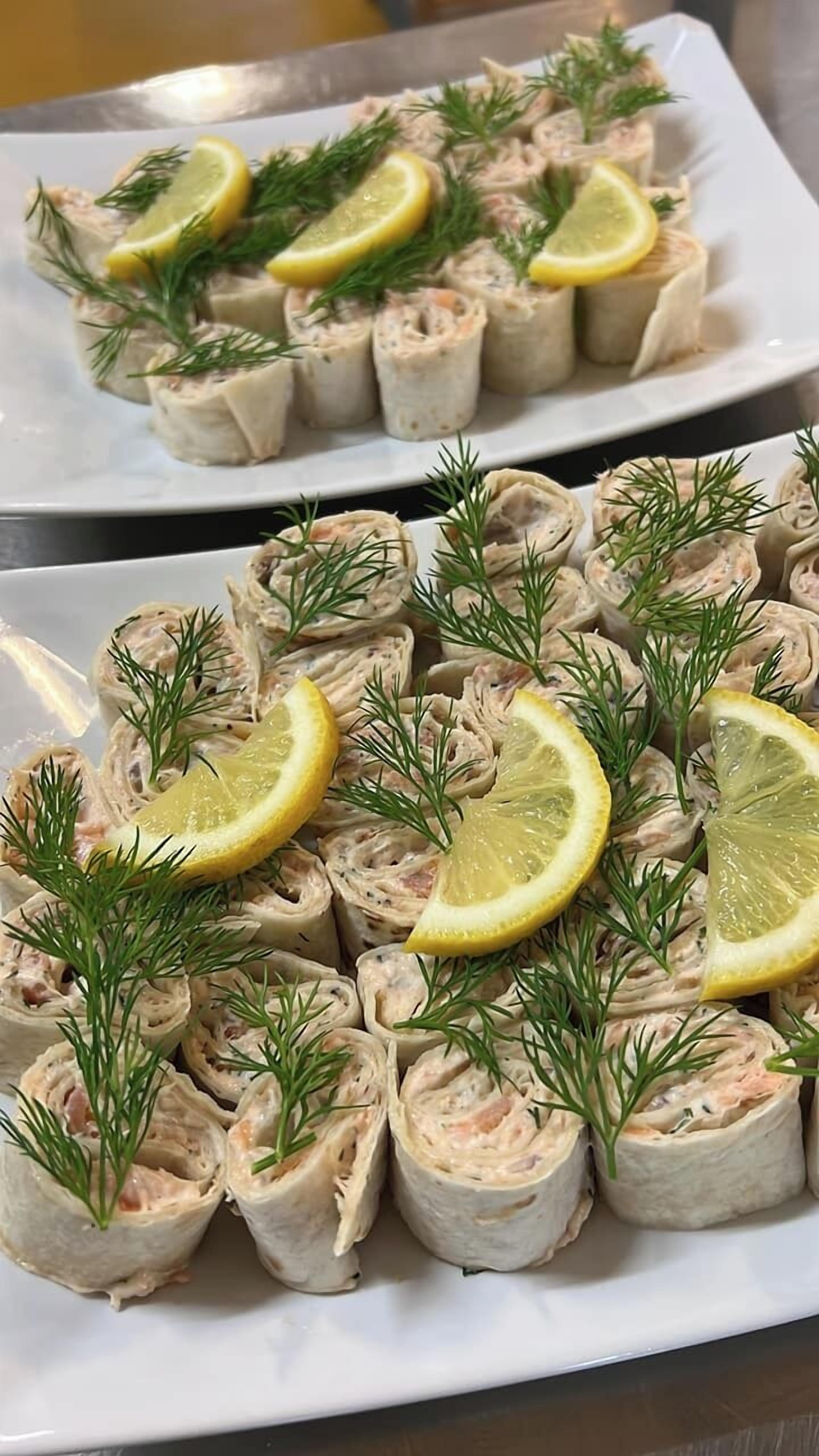 Dos platos de aperitivos enrollados, adornados con eneldo y rodajas de limón.