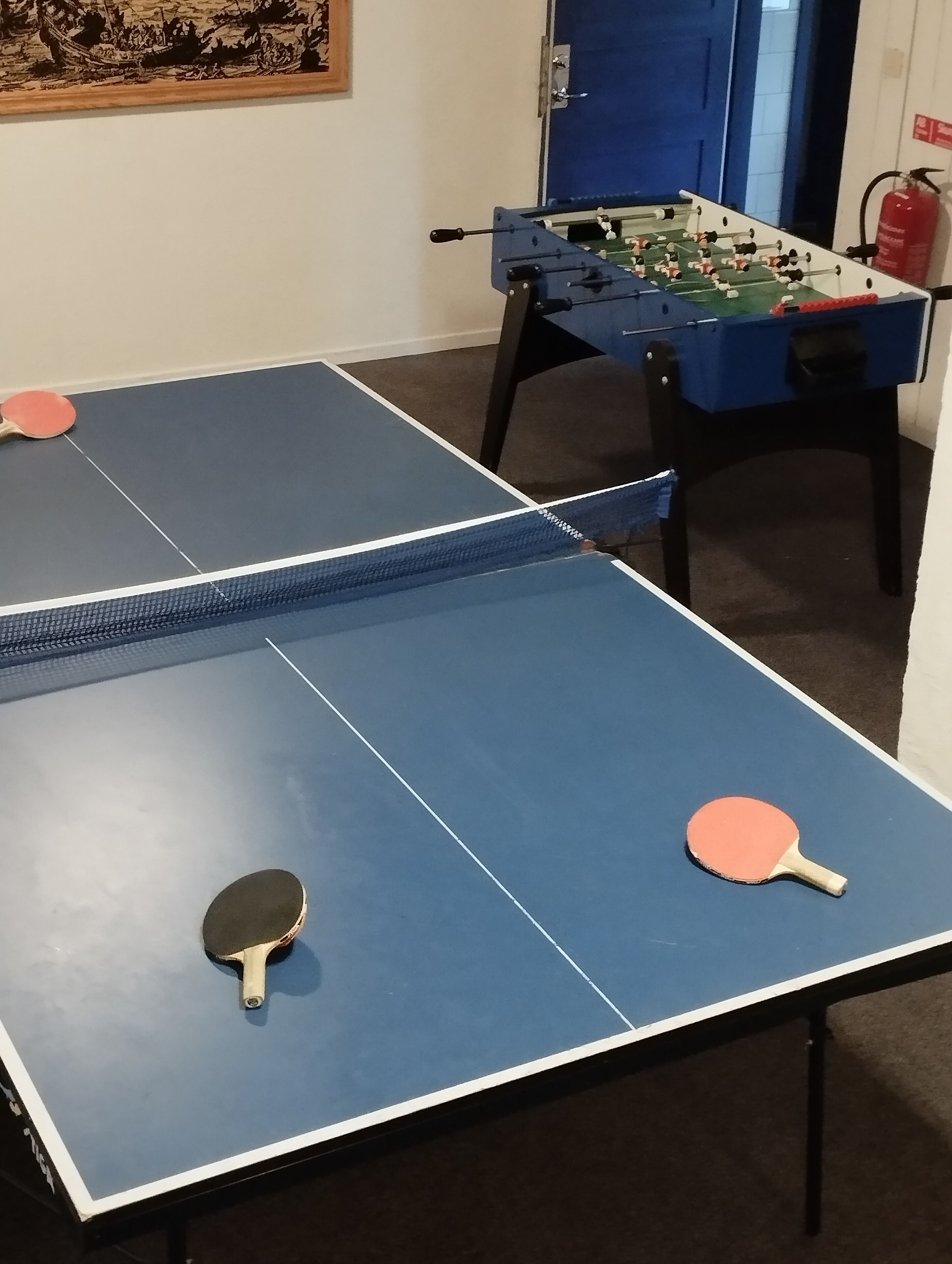 Una habitación con dos mesas de ping pong, una con palas a cada lado, y una mesa de futbolín al fondo.