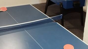 Una habitación con dos mesas de ping pong, una con palas a cada lado, y una mesa de futbolín al fondo.