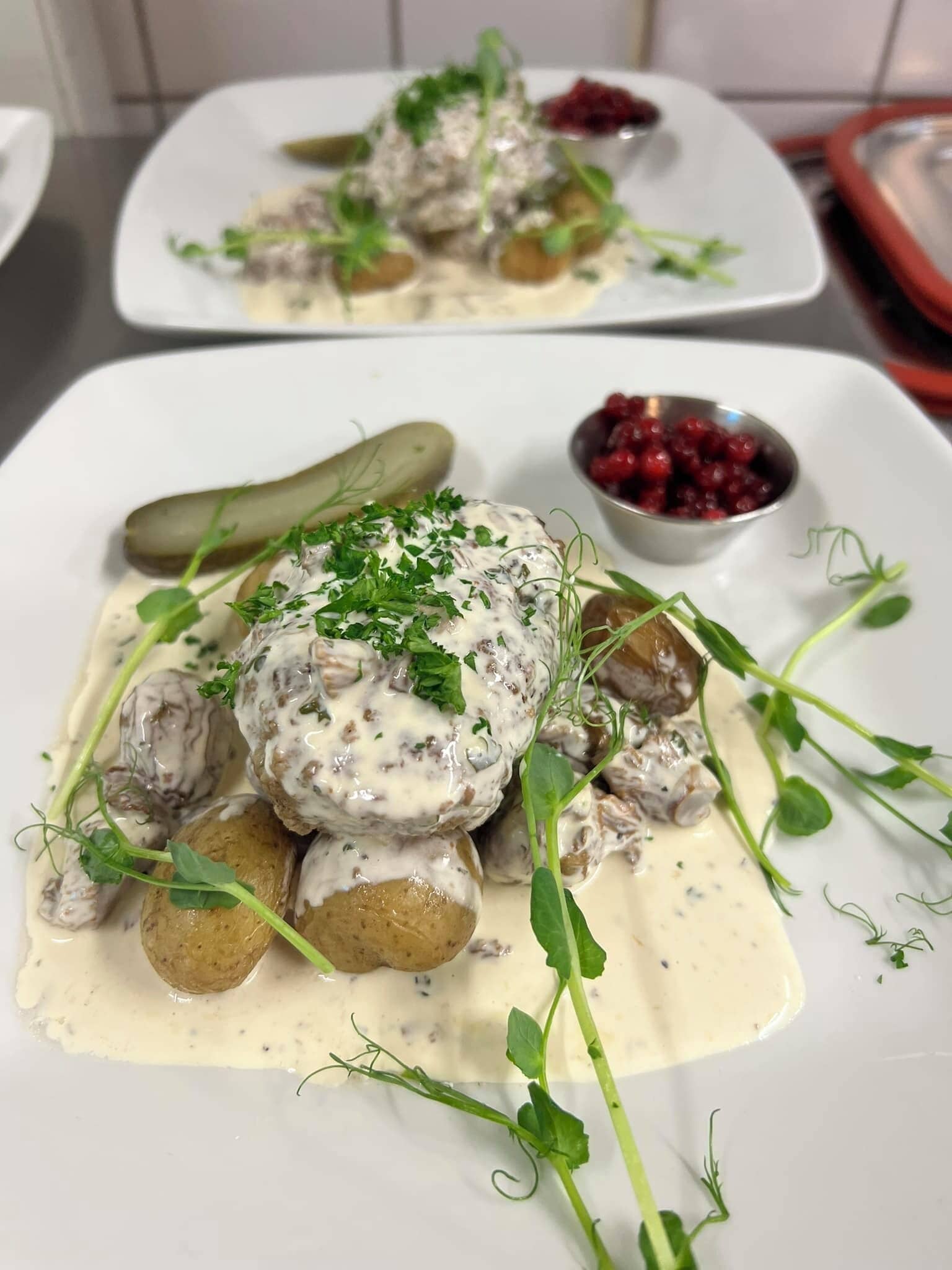Eine Portion schwedischer Hackbällchen mit cremiger Soße, garniert mit Kräutern, dazu Essiggurken und Preiselbeersoße.