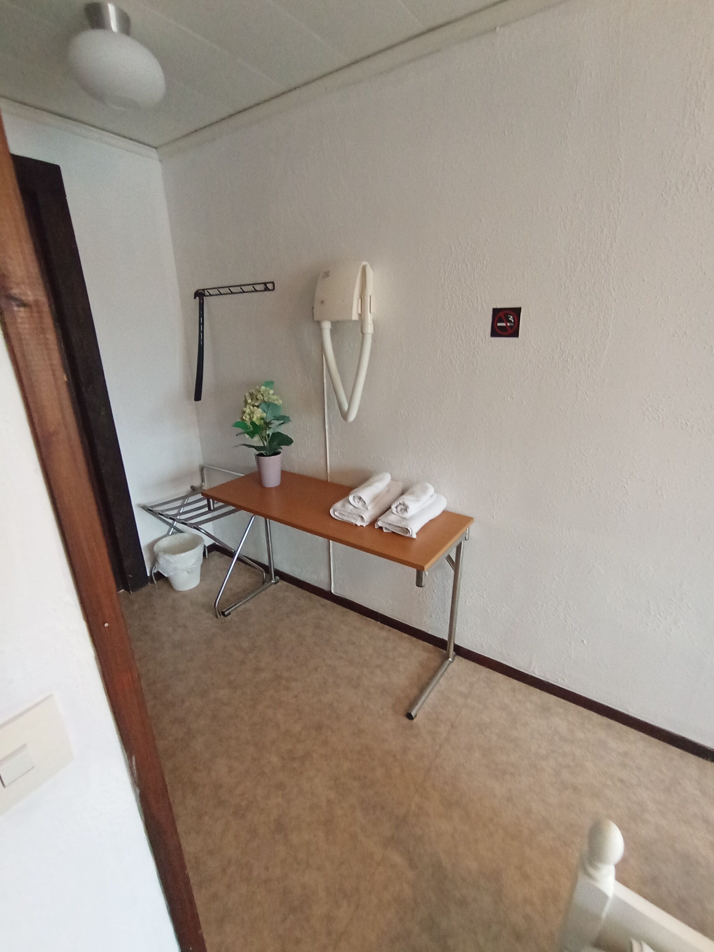 Una habitación con una mesa de madera, una planta en maceta, un perchero, un cubo blanco y un cartel de prohibido fumar en la pared.
