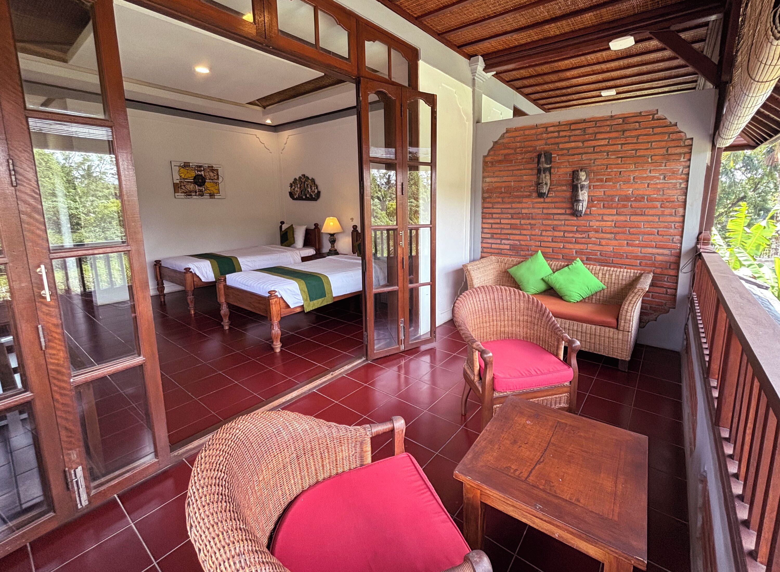 A room with two beds, a wicker sofa with green cushions, a wooden table, and a brick wall.