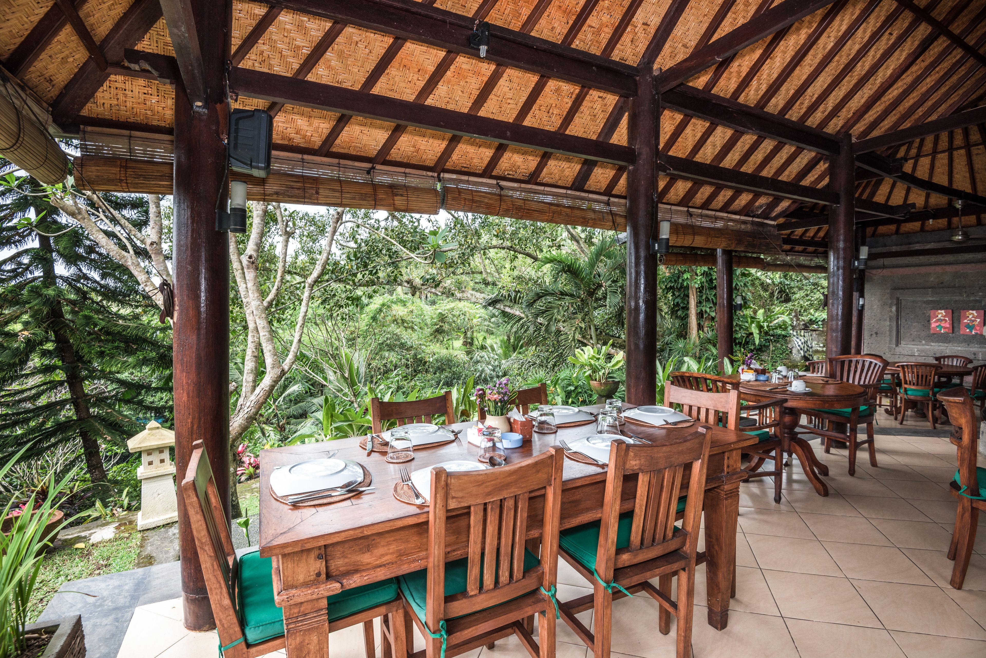 An outdoor dining area with wooden tables and chairs, surrounded by lush greenery and a thatched roof structure.