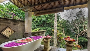 A stone bathtub filled with colorful flowers, a stone pedestal with a flower arrangement, and lush greenery in the background. - Hotel Bunga Permai (Ubud)