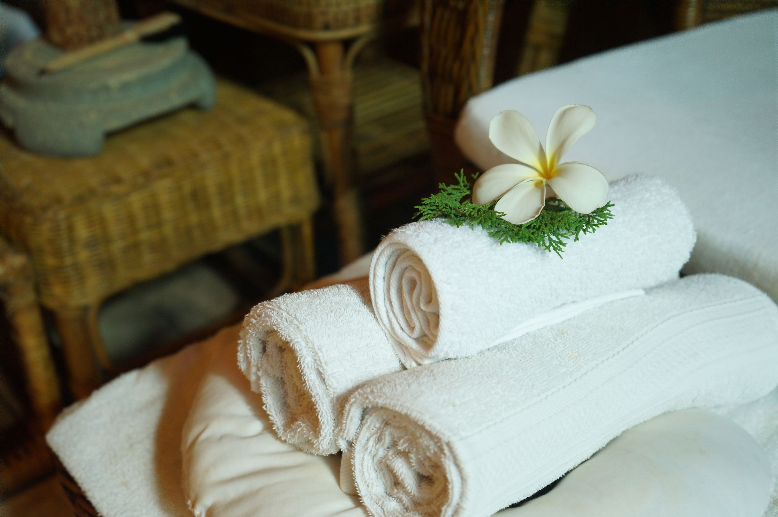 A stack of neatly folded white towels with a white flower and green foliage on top, placed on a bed in a room with wicker furniture.