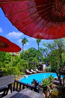 Zona de piscina con sillones reclinables y sombrillas, rodeada de exuberante vegetación y palmeras.