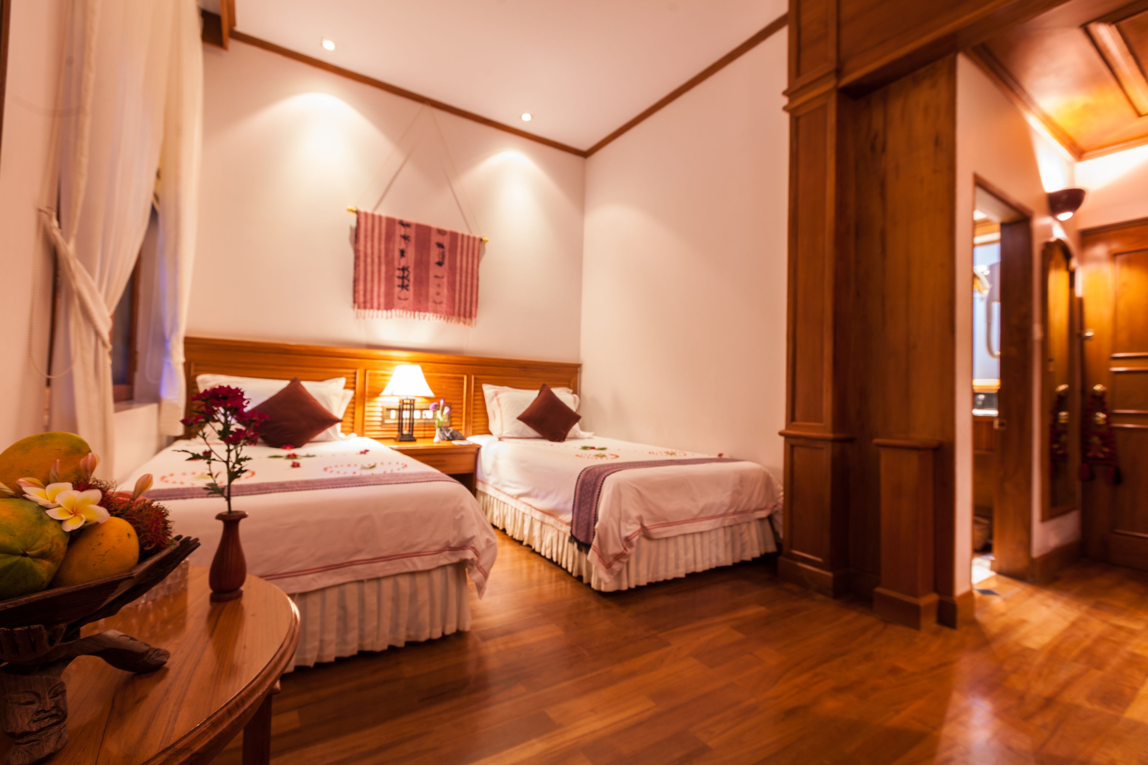 Habitación de hotel con dos camas, suelo de madera, una mesita de noche con lámpara, una pequeña mesa con fruta y flores, y un adorno colgante.