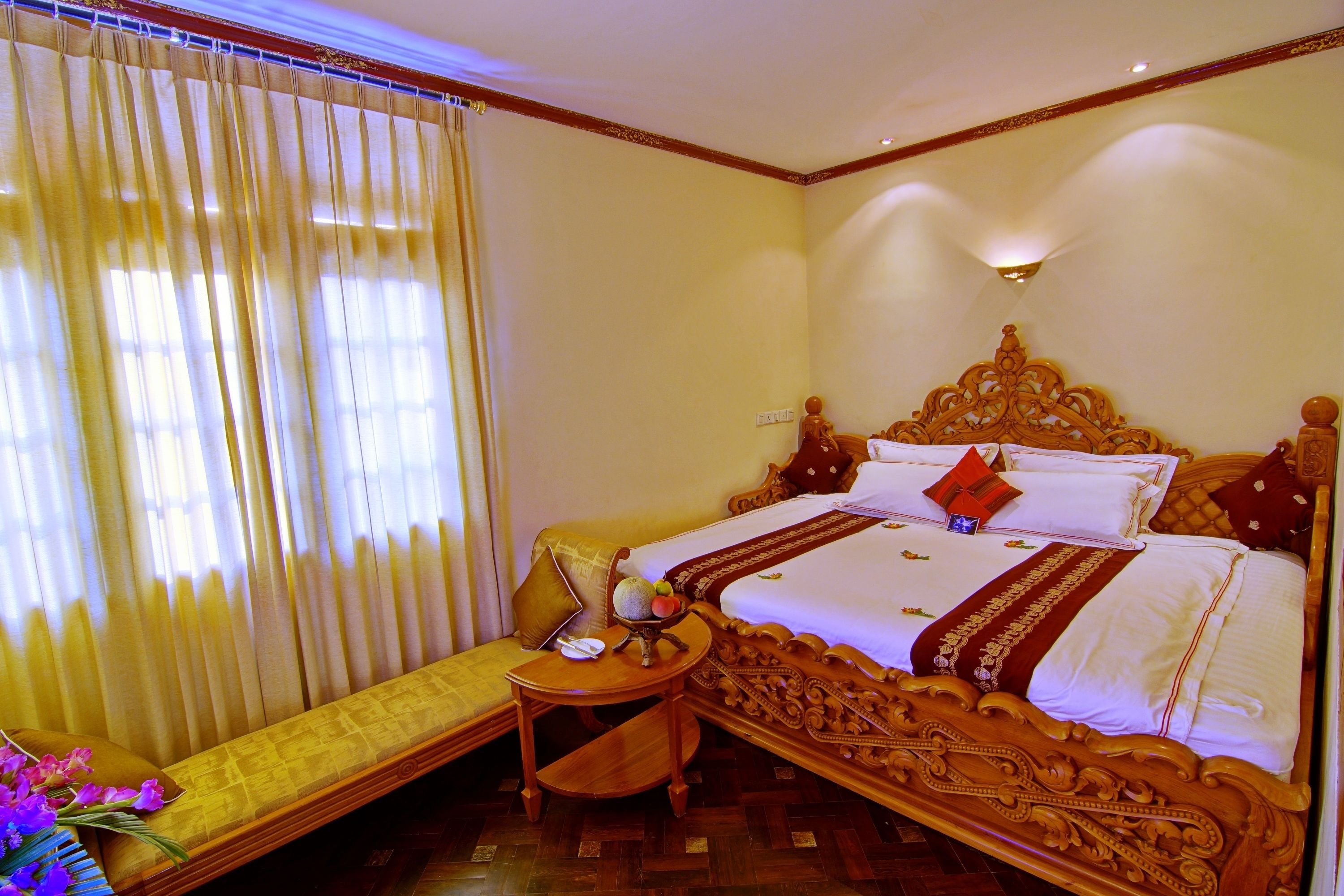 A hotel room with a large bed, a bedside table, a small table with a vase of flowers, and a bench with cushions.