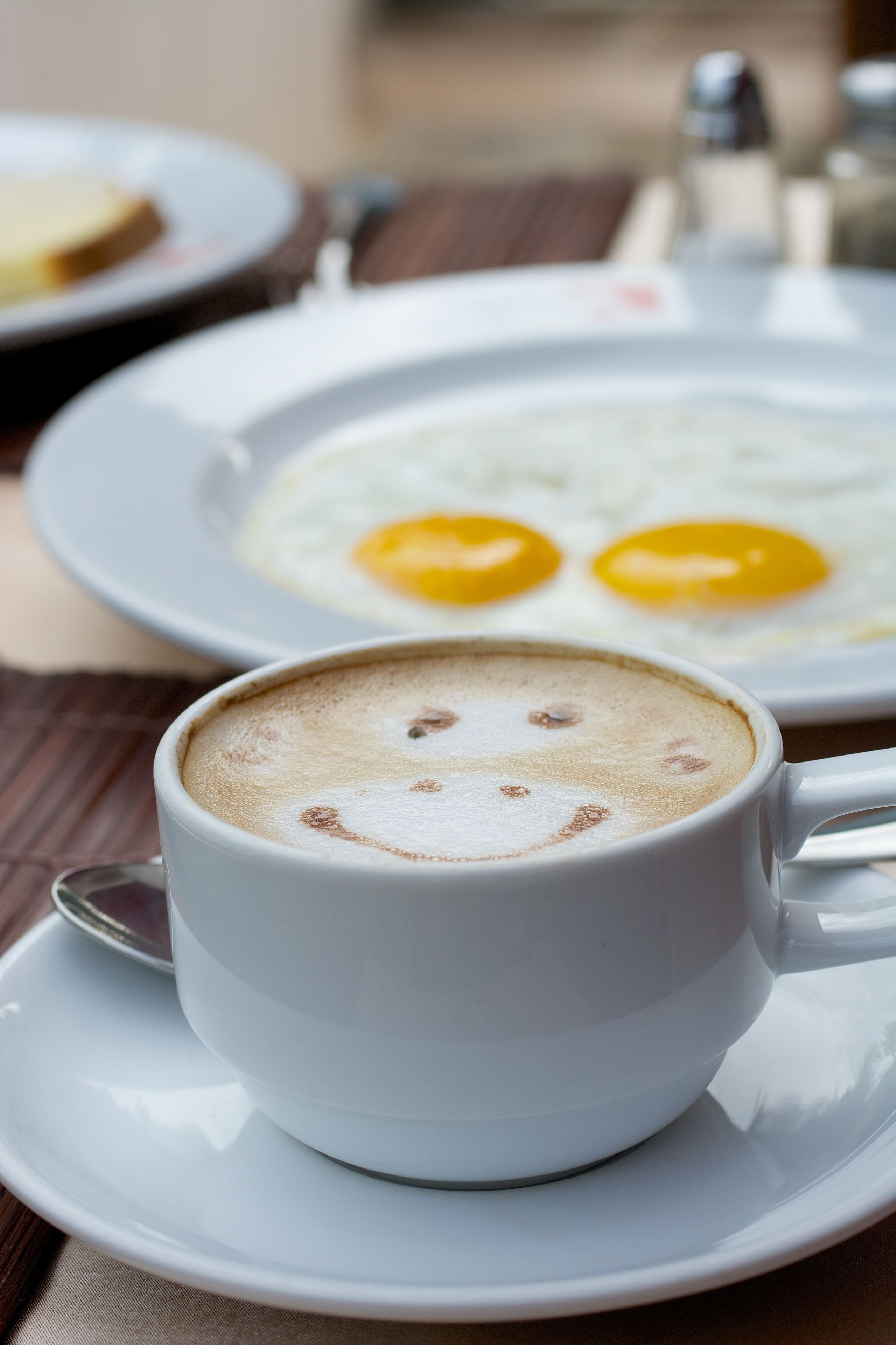 A cup of coffee with a smiley face on the foam, a plate of eggs, and a salt shaker on a wooden table.