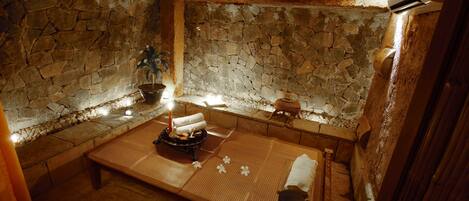 A stone-walled room with a tatami mat, a small table with a candle, and a potted plant.