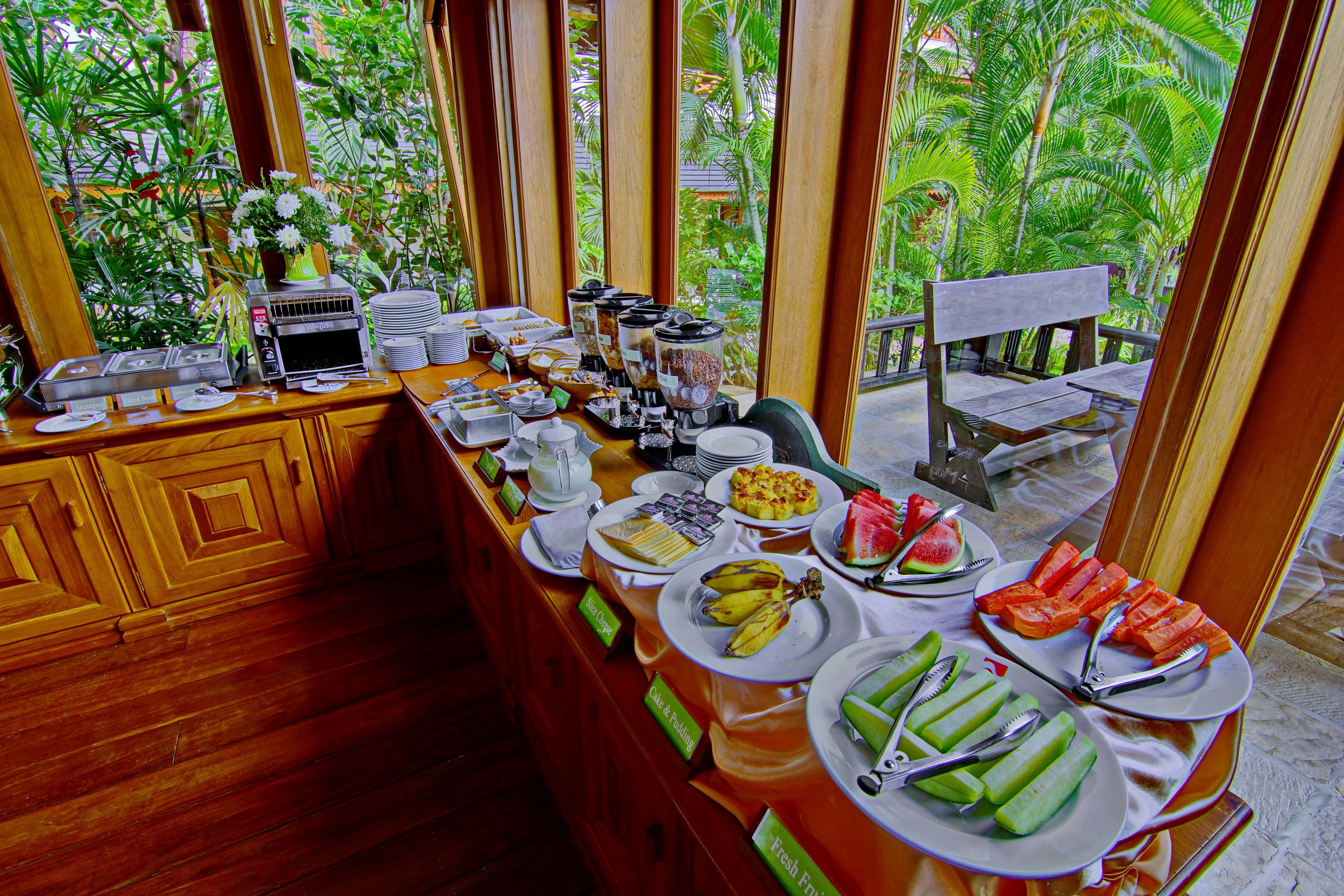 A buffet setup with various food items and a coffee machine, surrounded by wooden cabinets and large windows offering a view of lush greenery.