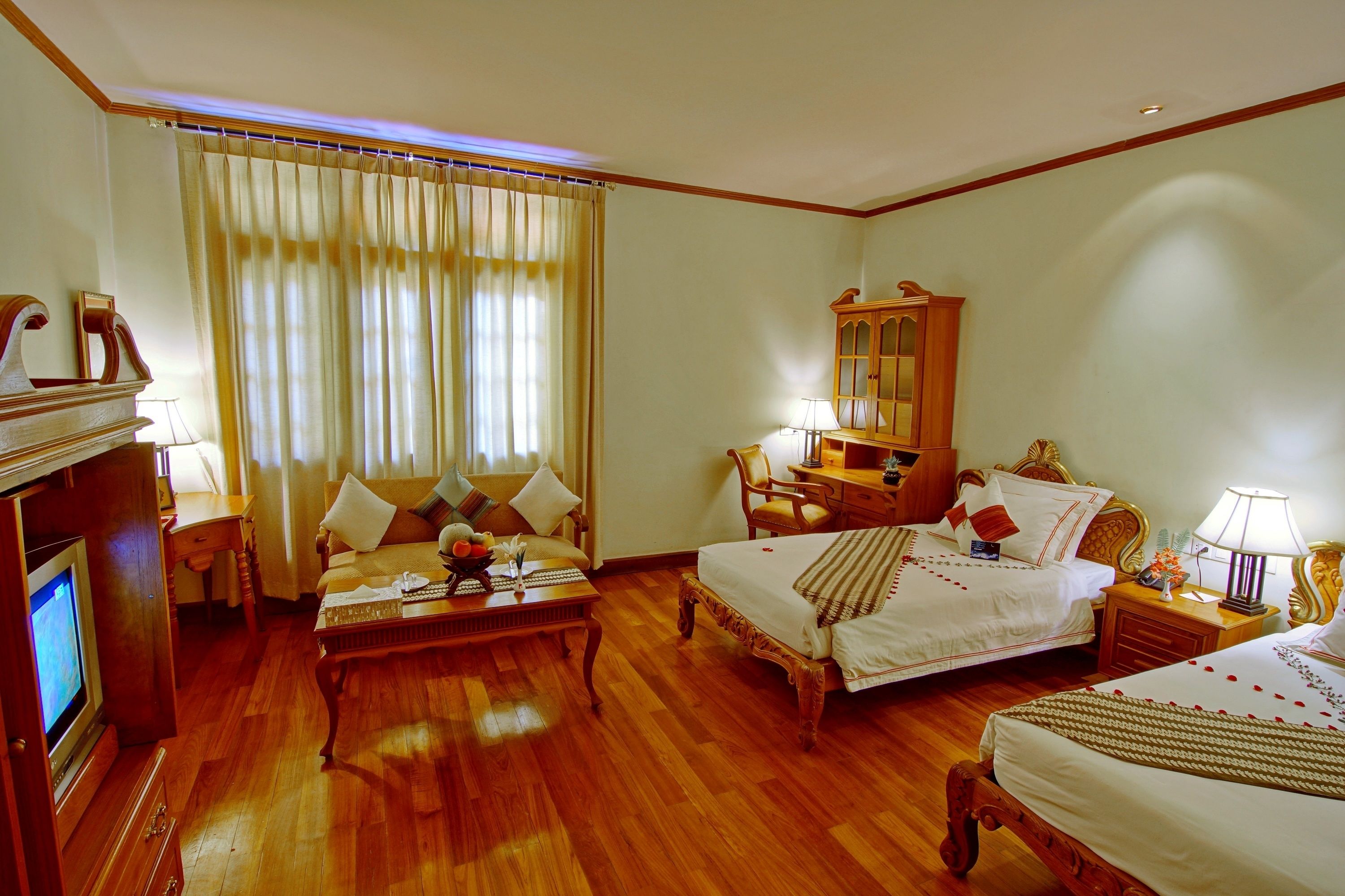 A hotel room with two beds, a wooden floor, a large window with curtains, a wooden cabinet, and a television.