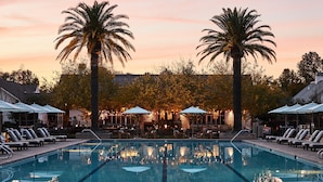 A swimming pool with lounge chairs and umbrellas, surrounded by palm trees and a restaurant area.