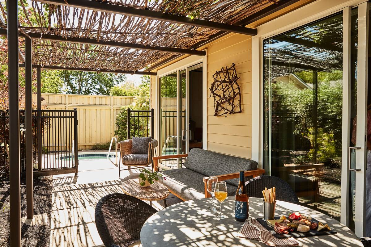 An outdoor patio with a pergola, seating, a table with food and drinks, and a view of a backyard.
