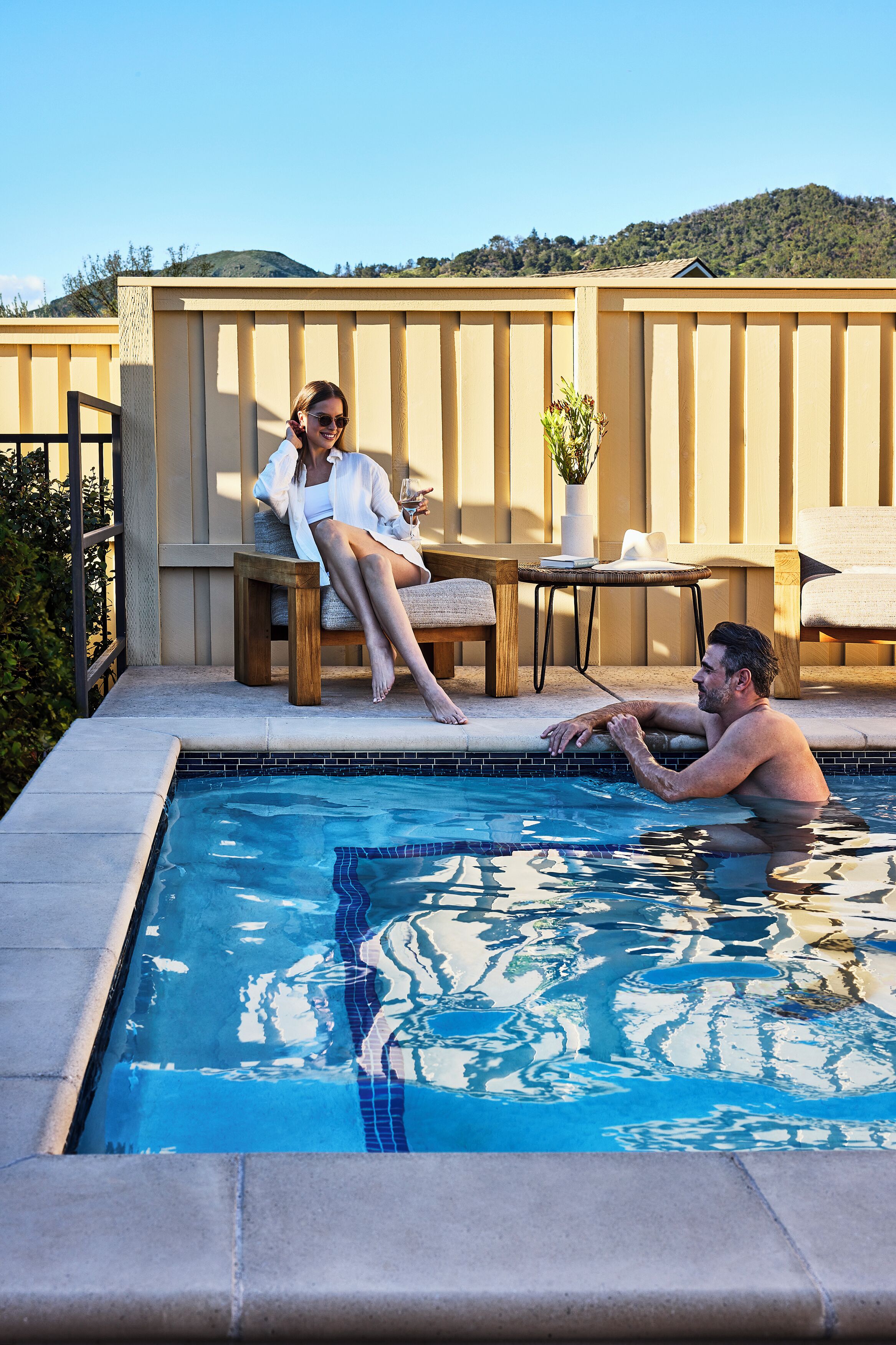 Zona de piscina con cubierta de madera, sillas de descanso y una mesa con planta en maceta.