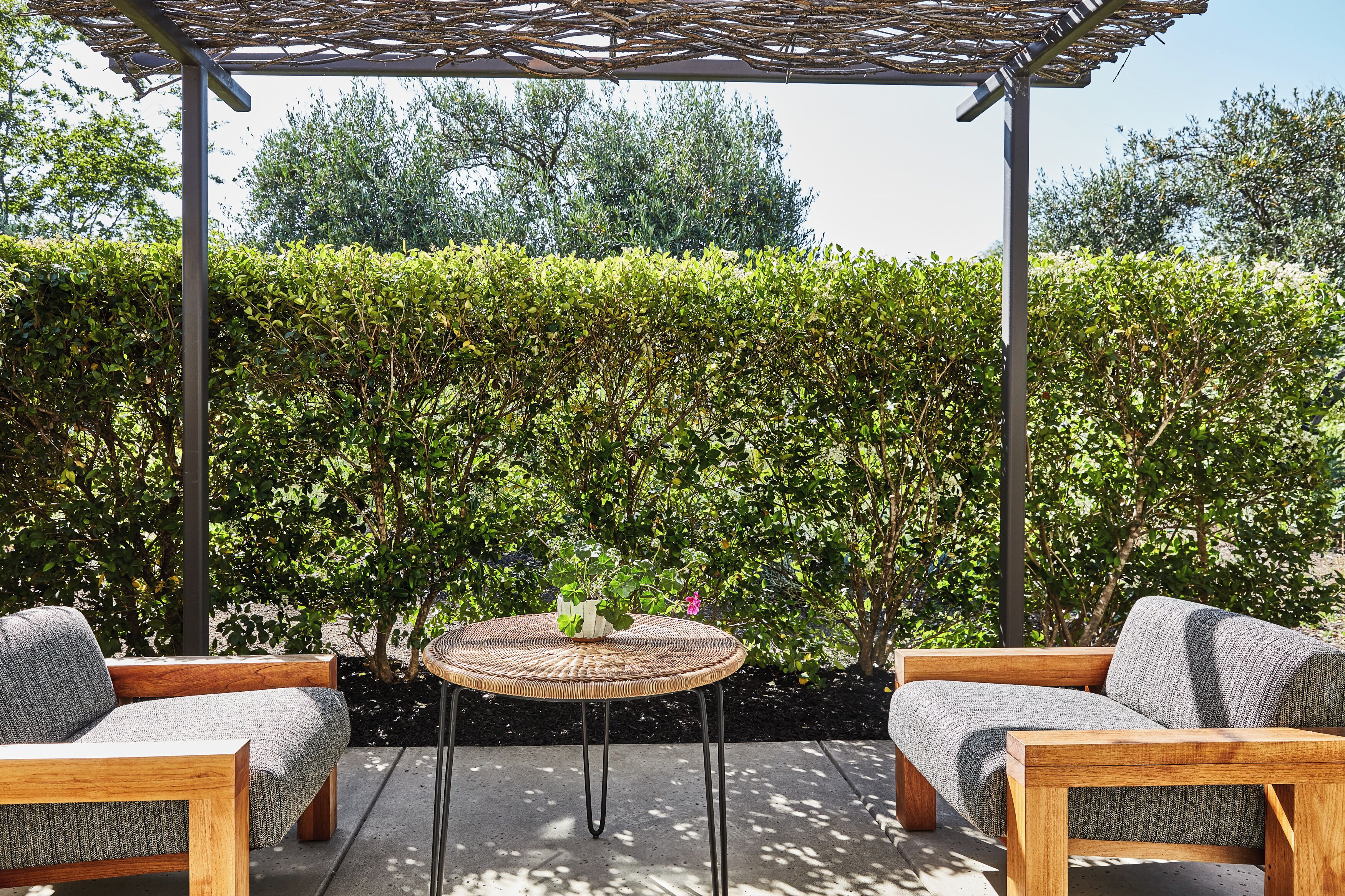 An outdoor seating area with a round table and two sofas under a pergola, surrounded by a well-maintained hedge.