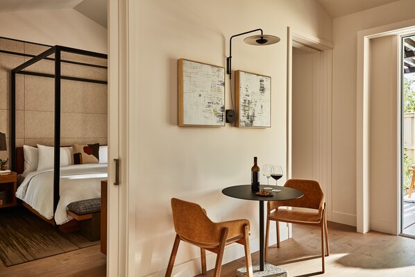A hotel room with a bed, bedside table, and a small round table with two chairs. There are framed artworks on the wall, a floor lamp, and a view of the outside.