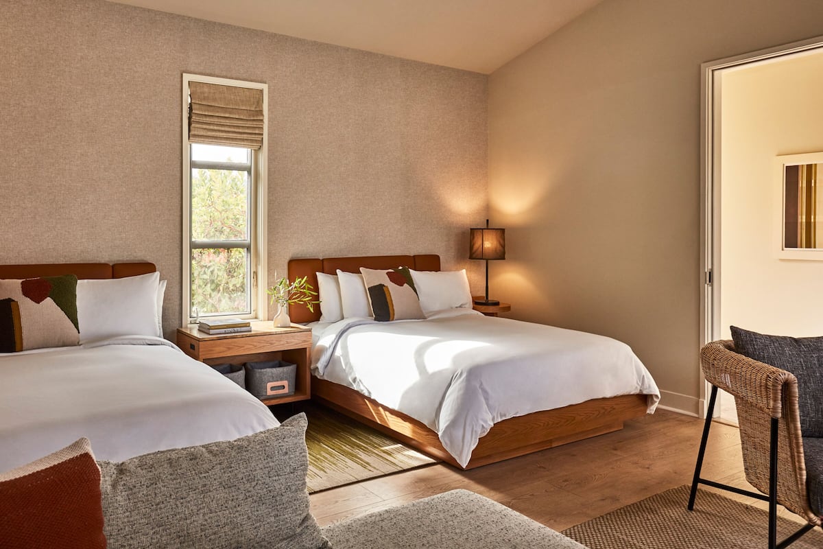 A hotel room with two beds, a window, a lamp, and a wicker chair.