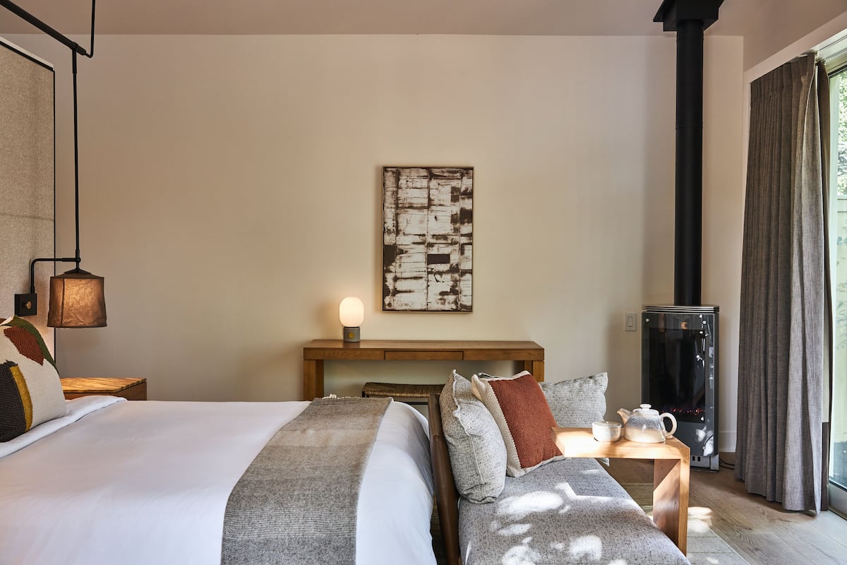 A hotel room with a bed, bedside table, lamp, artwork, and a heater.