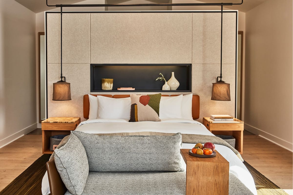 A modern hotel room with a large bed, two bedside tables, a nightstand, a sofa, and a wall-mounted shelf with decorative items.