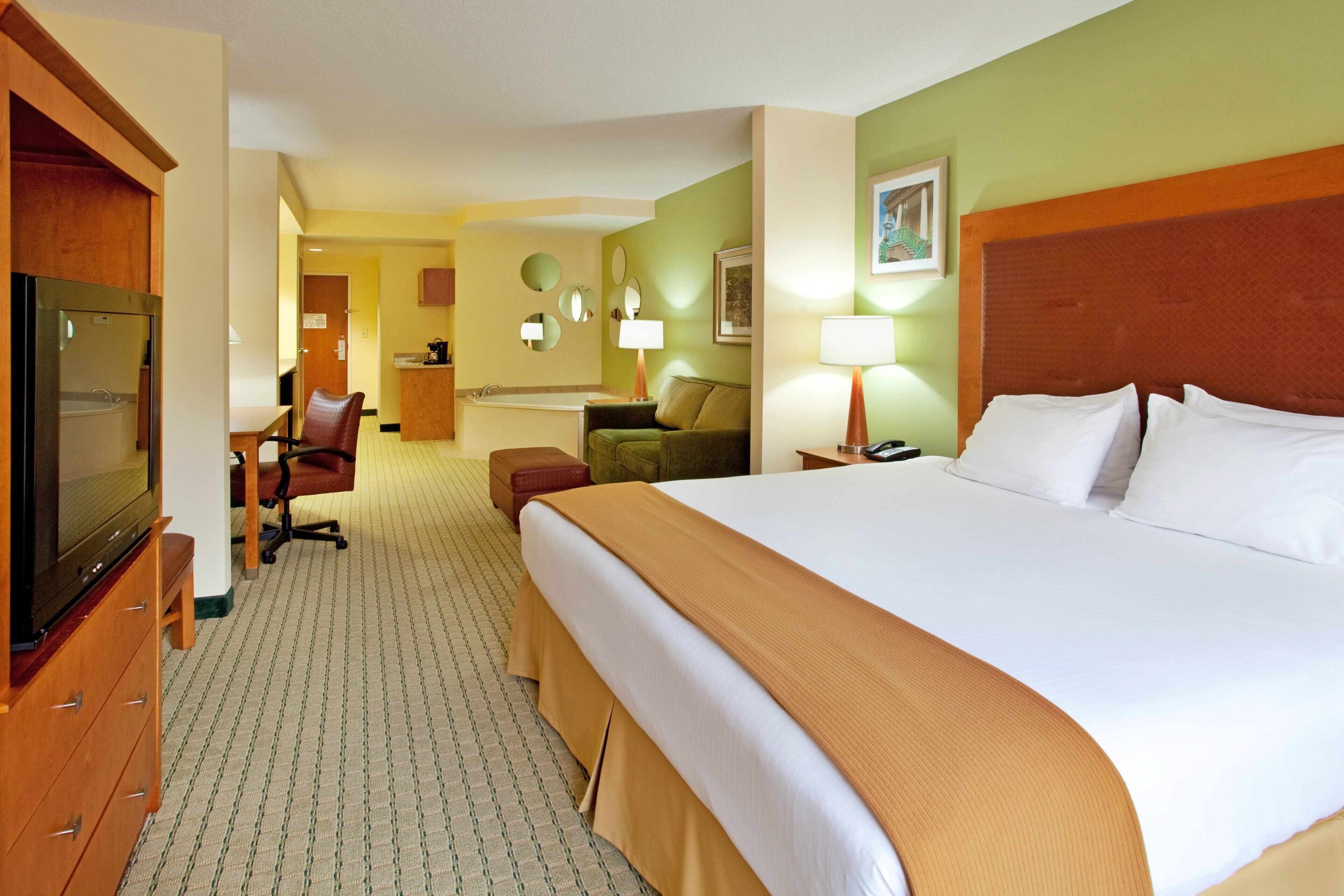 A hotel room with a large bed, a television, a desk with a chair, a sofa, and a small kitchenette.