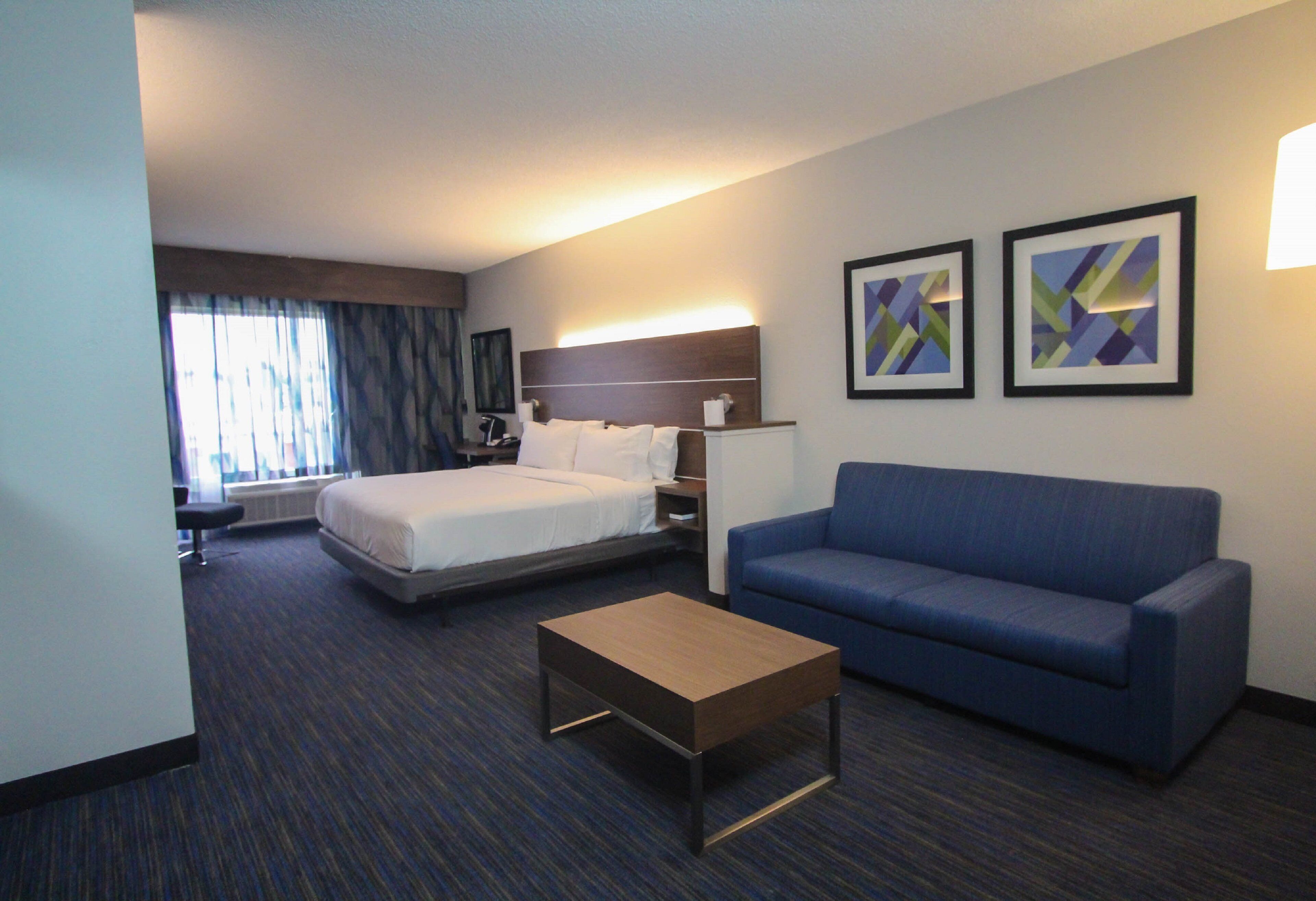 Una camera d'albergo con un letto, un divano blu, un tavolino in legno e due quadri astratti alle pareti.