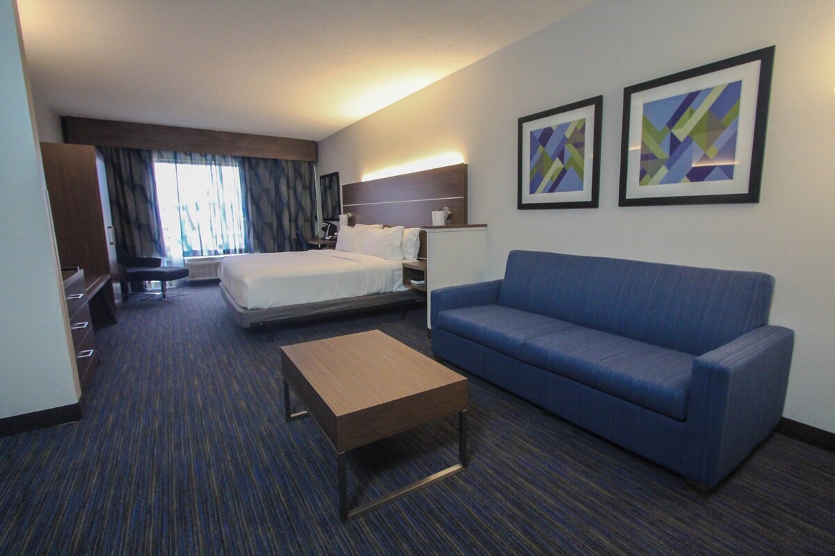 Une chambre d’hôtel avec un lit, un canapé bleu, une table basse en bois, un bureau et une chaise. Deux tableaux abstraits encadrés sont accrochés au mur.