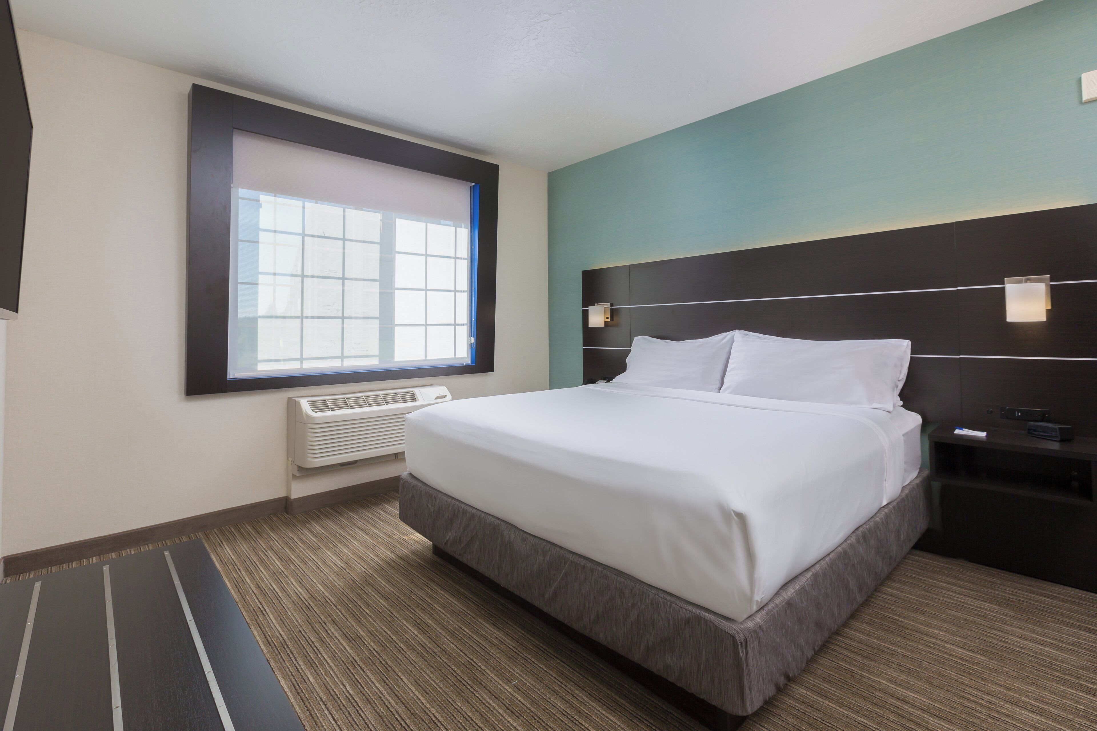Una habitación de hotel con una cama grande, una ventana con persianas, un calentador en la pared y una mesita de noche con lámpara.