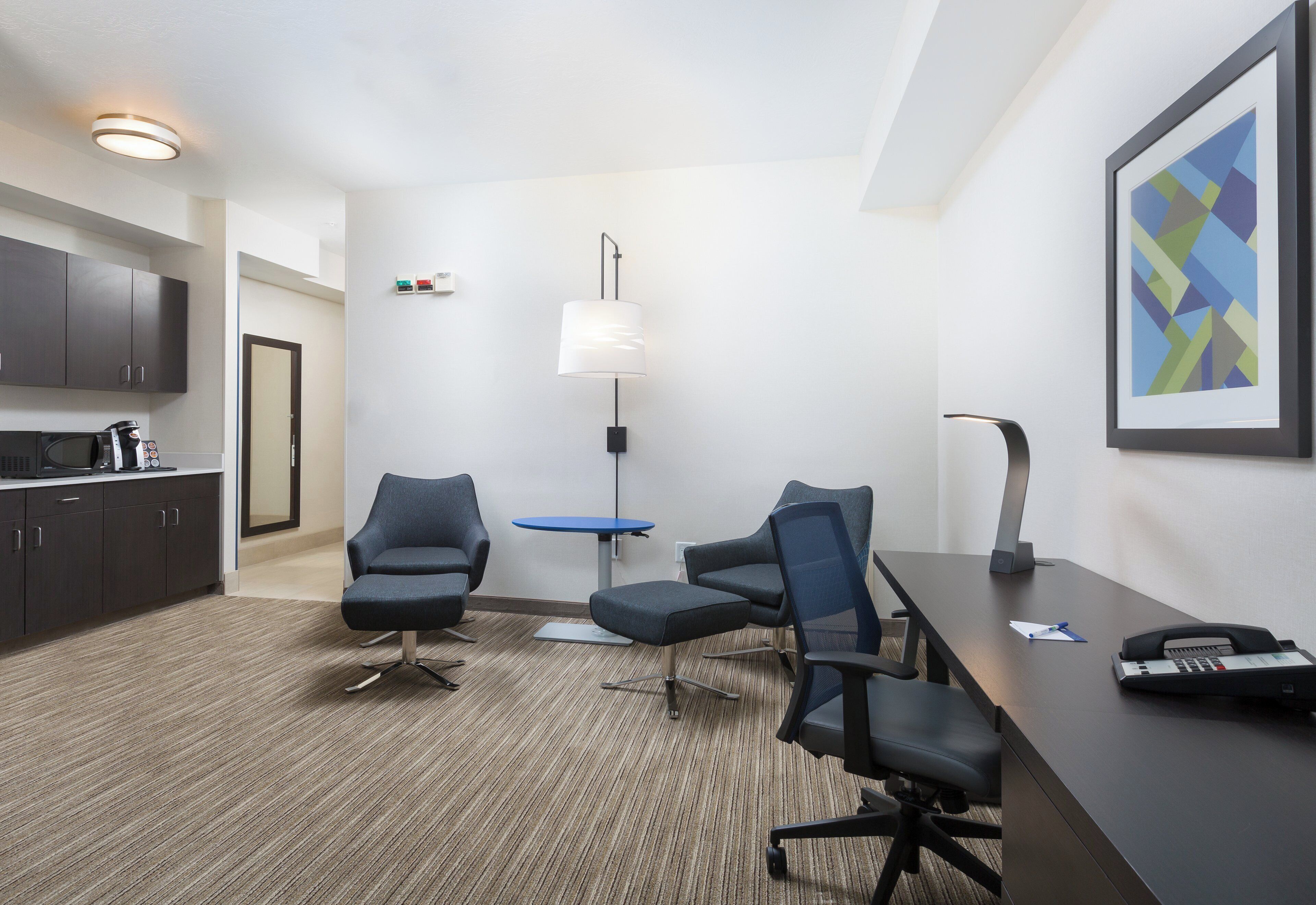 A modern hotel room with a kitchenette, two chairs, a desk, a lamp, and a framed abstract painting.