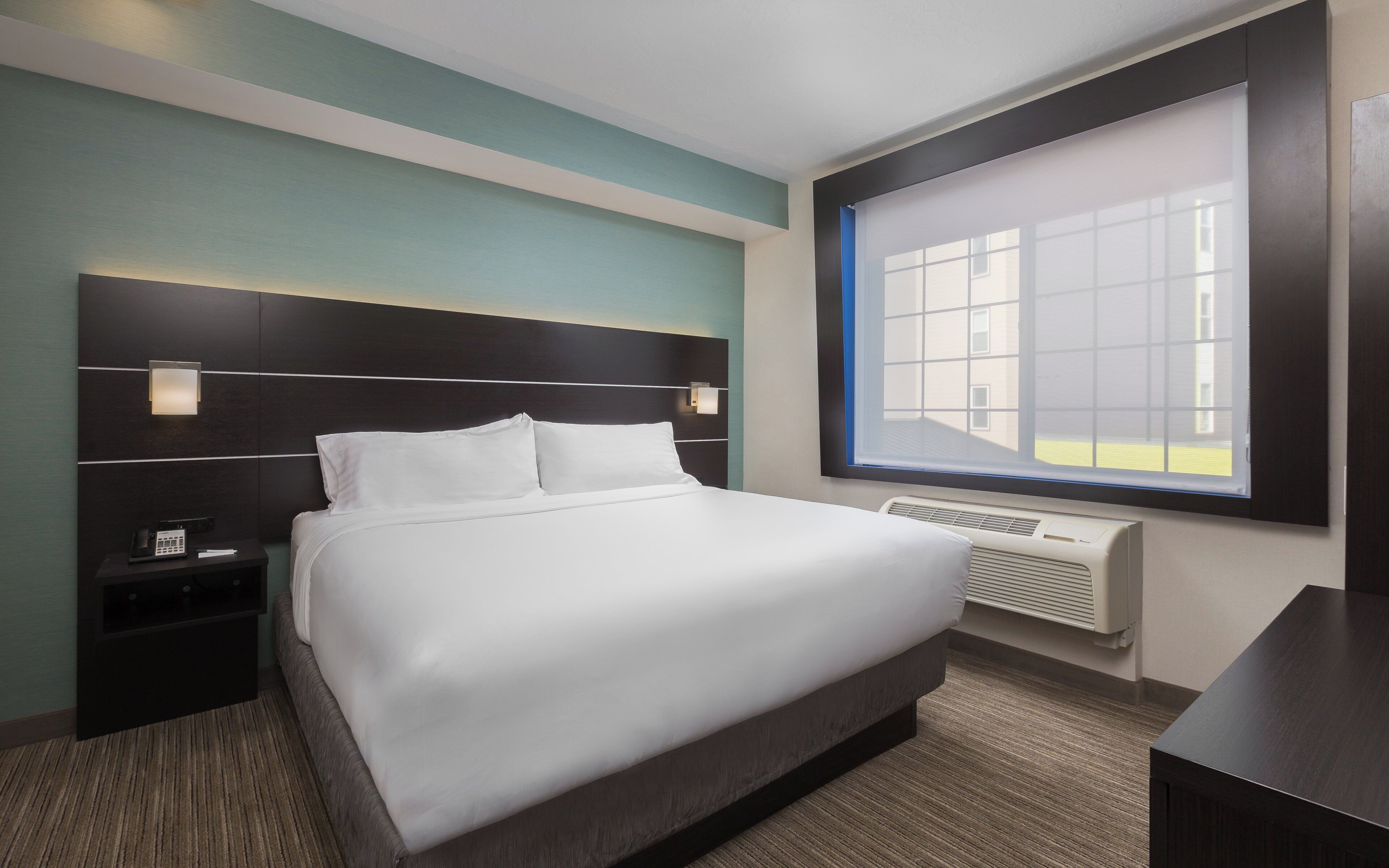 A hotel room with a large bed, bedside tables, a window with a view, and a built-in air conditioning unit.