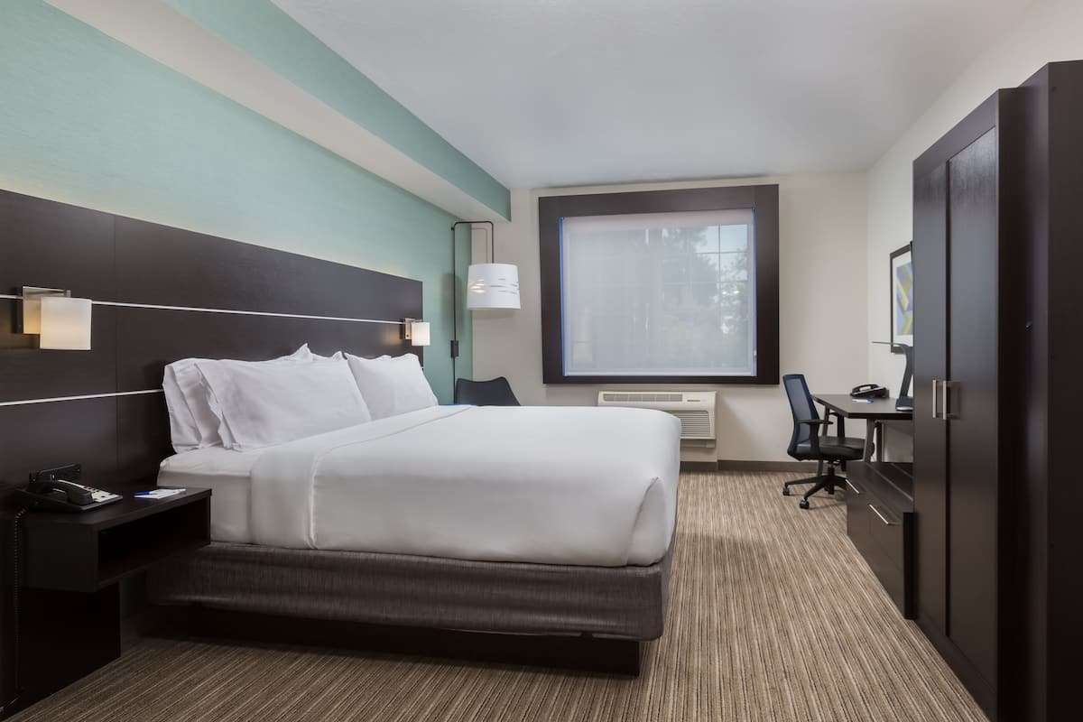 A hotel room with a large bed, a desk with a chair, a window with blinds, and a wardrobe.