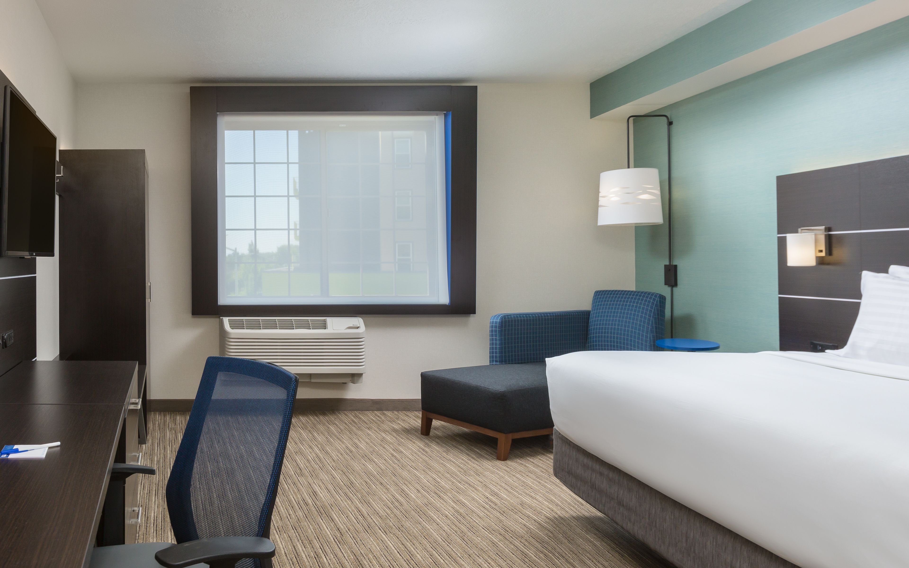 A hotel room with a bed, a desk, a chair, a small table, a window, and a wall-mounted lamp.